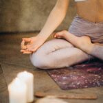 A woman practicing yoga indoors in lotus pose with candles creating a calming atmosphere.